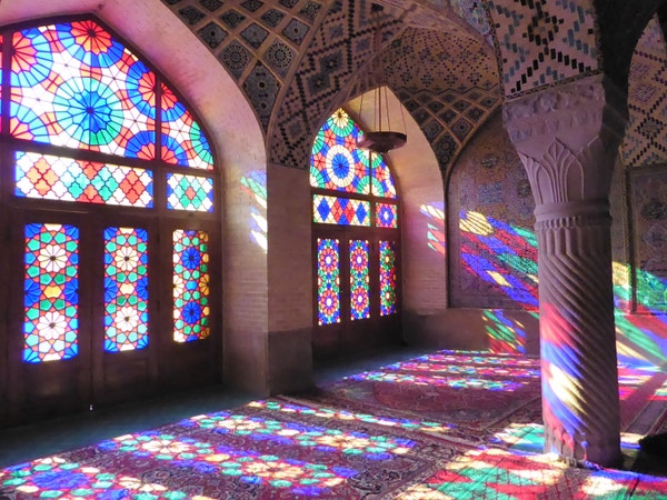 Interior view of a mosque. Bright sunlight comes from the left, through large arched stained glass windows. The light falls onto a floor covered in red carpets. The whole scene is suffused with pink, yellow and blue light.