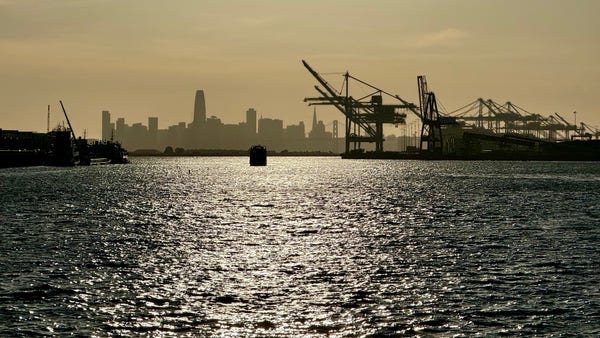 San Francisco skyline and port of Oakland cranes silhouetted by hazy sunset sky. 