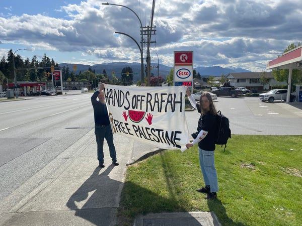 Two people standing with a large banner that says "Hands of Rafah and "Free Palestine". They are pointing toward a roadway from a sidewalk and grassy area beside a gas station.
