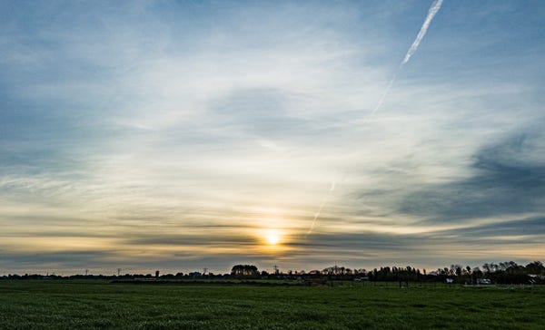 The sun rising through layers of cloud near the eastern horizon, looking towards Leiden from the road between Wassenaar & Voorschoten. There is a rather fuzzy 22° halo around the sun, a contrail to the upper right, & a flat green field in the foreground.