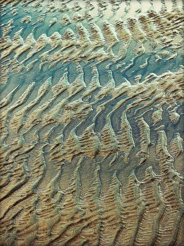 A colour photograph of waterlogged sand in a pattern similar to dogtooth. The water over the sand reflects some of the blue sky above, contrasting the sand.