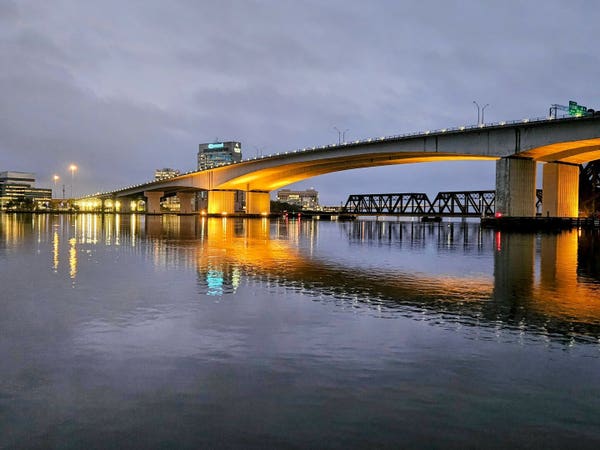 Beneath stormy skies a large concrete bridge spans a river with bright yellow lighting illuminating the underside of the bridge, with colorful shoreline lighting, all reflecting upon the water below.