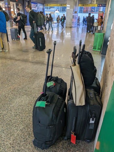 Four small roller bags of carry on size, with a small backpack purse stacked on top. The shot is taken inside the train station and other passengers are in the background.
