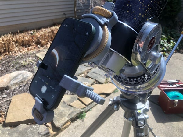 A telescope attached to a tripod with a smartphone mounted on the eyepiece with a silver 3D printed mount, capturing an image of a partial solar eclipse.