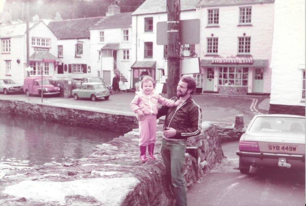 Photo taken about 40 years ago of my Dad supporting little me on a wall next to water with white buildings behind us
