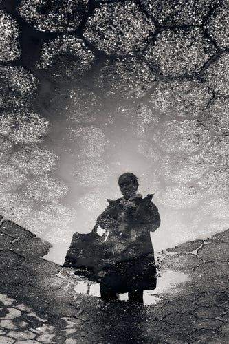A black and white image of a person reflected in a puddle.

The puddle is situated on a tiled hexagonal stone ground. The tiles are split between lighter and darker variants. The darker ones near the bottom create an edge in the middle, right around the reflected person's waist. The lighter tiles above are mixed with dirt and grime, almost like a cloudy sky.

The person is has long dark hair pulled back in a bun or ponytail. They're wearing a dark coat and white scarf. They have a large tote resting in the crook of their right arm. They are holding something in their hand and looking down at it with a furrowed brow. It's not clear what.