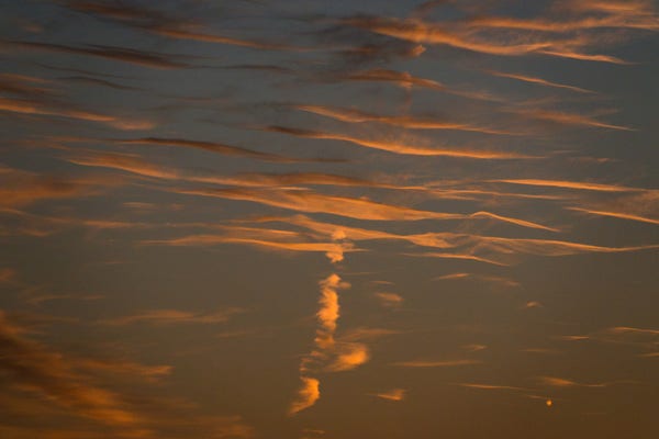 Horizontal wisps of rosy sunset clouds against a darkening dusk sky. The remains of a jet stream is crossing them vertically, and towards the top, the shadow of night is falling on the clouds.