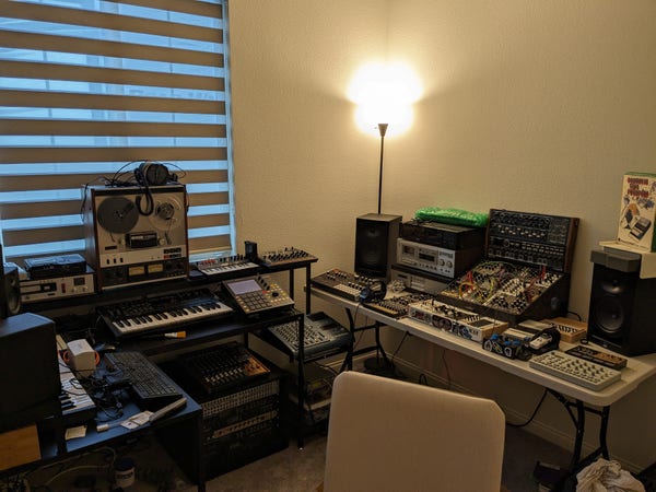 A picture of a home music studio based around synthesizers and tape recorders. A wide variety of gear is visible including a reel-to-reel tape recorder, multiple cassette decks, a medium-sized modular synthesizer (12u, ~350hp total), drum machines, rack-mounted effects and more.
