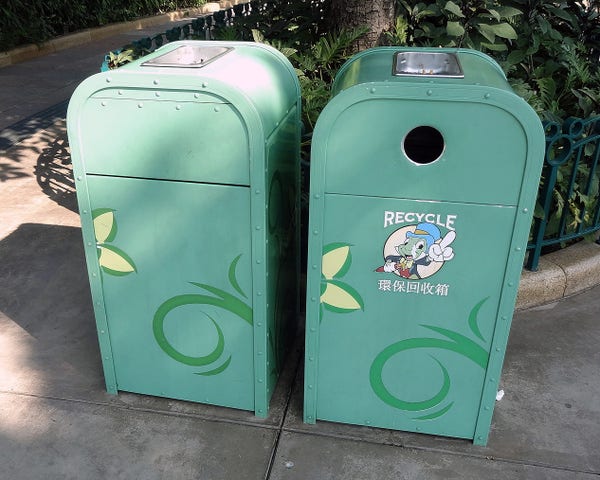   Two green trash cans with decorative leaf patterns and a cartoon character on the front, located outdoors near a fence and foliage. 