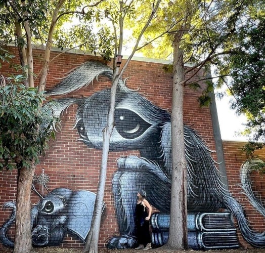 Streetartwall. Street art wall. A mural with two fluffy fantasy figures has been painted on a brown brick wall in a park. A small gray-black, fluffy creature with big eyes, curved ears, a broad nose and two tufts of plants on its head stands there with a book in its hand. It looks like a mixture of squirrel, mouse and rabbit. It is standing in front of a larger animal with a similar appearance, which is sitting on a pile of books. (There are some trees in front of the mural and the young artist in black dungarees is looking up at her fantasy friends).
Info: The mural is part of a larger picture. Another fluffy friend is standing to the left of it. (extra photo)