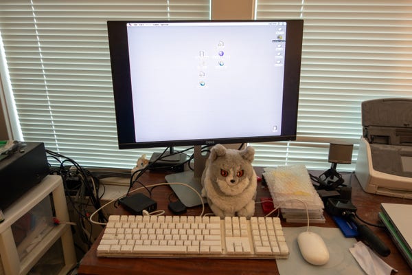 Computer monitor showing a Mac OS 7 desktop with an Apple Keyboard and Mouse and a Lailaps plush.