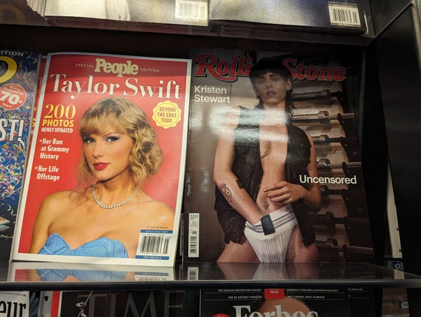 A magazine rack featuring a dolled up Taylor Swift in baby blue dress and pearls, and a butch af Kristen Stewart in a black leather vest and jockstrap 
