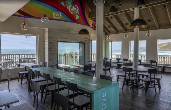A restaurant dining room with a natural wood floor and open-beam ceiling. The chairs are of modern design and the tables resemble stained barn wood. On one wall is a colorful mural above a series of windows looking onto a dining patio; beyond is a sandy beach, rocky cliffs covered with scrub, and the breakers of the Pacific Ocean