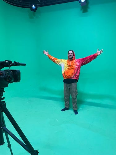 Musical artist Dizraeli in a tie dyed sweatshirt and khaki jeans, with his arms up and smiling. He is on a green screen with a film camera in front of him.