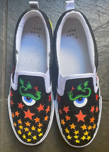 A pair of canvas shoes painted with stars and eyeballs with snakes as eyebrows and leaves on the ends of the snakes's tails.