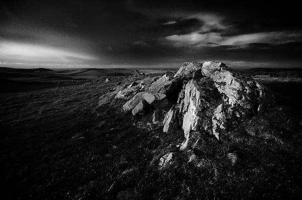 Atmospheric black and white photo of rocks protruding from a grassy field, under a dark sky.