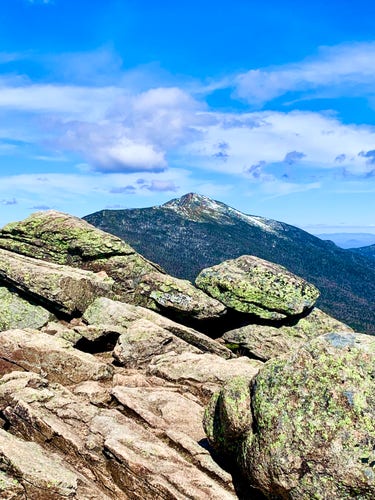 Photo of distant snow-covered mountain peak as viewed from another peak.  Boulders in foreground, sky is blue with puffy white clouds. 

Photo taken last month from Mt Liberty, White Mountains, New Hampshire when I snowshoed to the summit. The distant peak is in the direction of Mt Lafayette.