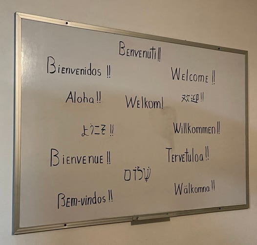 Photo of a whiteboard with the phrase “Welcome!!” written in 12 different languages (English, Italian, Spanish, Portuguese, Finnish, Hebrew, et cetera), but the Hawaiian one is “Aloha!!”