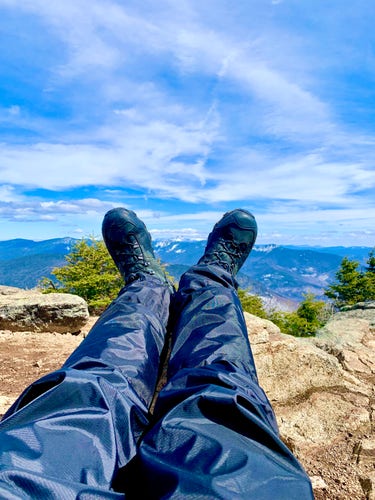 Photo of my legs and feet outstretched in front of me while lying down and view of distant mountains seen on horizon.  Mountain peak is viewed between my feet. Photo taken at mountain summit with full panoramic view.  Camera near waist aimed along length of legs toward feet.  I am wearing thick black rain pants and black winter boots.   I am lying on gray rock slabs, with short evergreen trees at mid-range and mountains at far range.  Sky is blue with white clouds.

Mt. Liberty, White Mountains, New Hampshire.