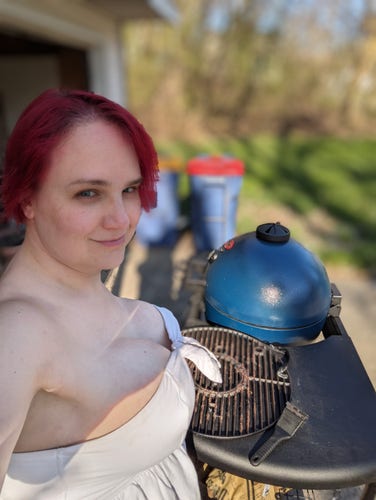 The poster stands in front of a blue kamado grill outside. She's wearing a strapless white dress that shows a lot of cleavage. 
