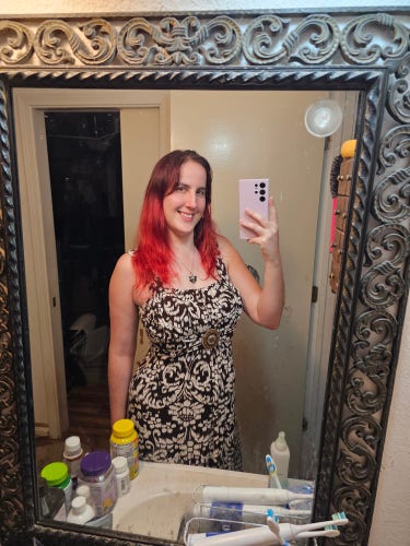 Photo of me with red hair smiling in the mirror while wearing a sleeveless brown and white dress synched at the waist with a beaded center medallion 