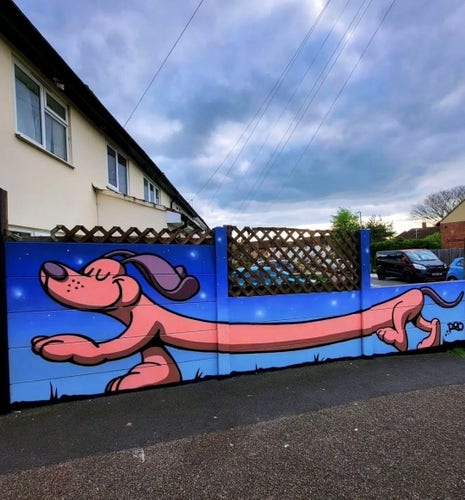 Streetartwall. A cheerful mural of a very long dachshund was sprayed on a low street wall. It looks a bit like a long pink sausage with legs and long floppy ears. The extra-long, good-humored animal trudges through a nocturnal scene with a blue starry sky and spreads good cheer.
(The photo shows a wooden lattice over the wall and behind it a one-storey long house and a gray cloudy sky)