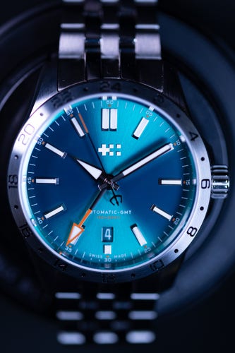 A close-up photo of a stainless steel Christopher Ward watch, the dial is a gorgeous teal/blue/turquiose colour, it's a GMT watch with an orange GMT hand.