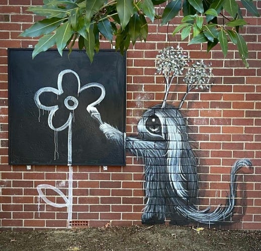 Streetartwall. A mural with a fluffy fantasy figure was sprayed/painted on a brown brick wall in a park. A small gray-black, fluffy creature with big eyes, curved ears, a broad nose and two tufts of plants on its 2head stands there with a paintbrush in its hand. It looks like a mixture of squirrel, mouse and rabbit. It is drawing a white flower, like a child's drawing, on a black board and the stem on the wall below. (Green leaves hang into the picture from the top right.
Info: The mural is part of a larger picture on this wall. There are two more fluffy creatures on the right-hand side.(Extra-Photo Part 2/2)
