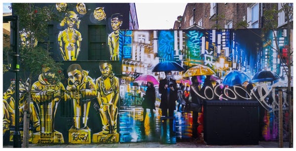 A vibrant street mural featuring portraits of individuals and abstract urban scenes, with pedestrians walking in front of the artwork, some carrying umbrellas.