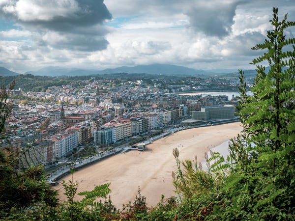 View from a mountain through the trees down onto a town with a big beach going towards the sea to the right. The houses are colourful and the weather cloudy, but with breaks for sun rays