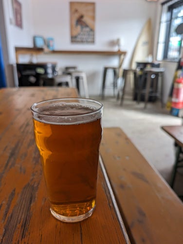 A pint of amber-coloured ale in a pint glass on a wooden table. In the background, blurred, there is small area with a wooden bar against a white wall and a few stools, all lit by natural daylight from a window on the right.