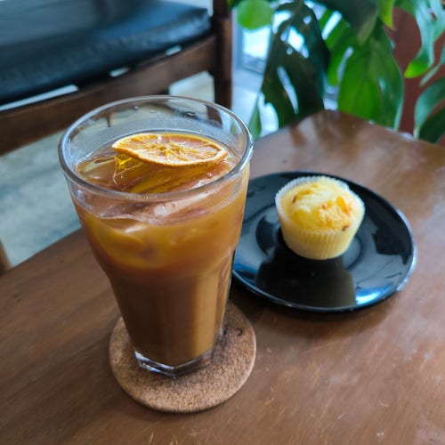 Tall glass of iced coffee with an orange slice on top and a yellow muffin on a black plate in the background