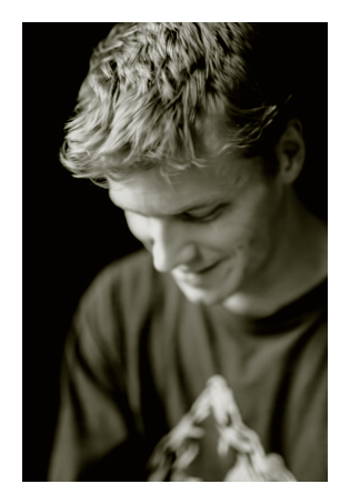close up. head and shoulders of a mid-20s guy with shorter, wavy light hair. he's wearing a dark tee with mountain peak graphic. he's looking down, smiling.
