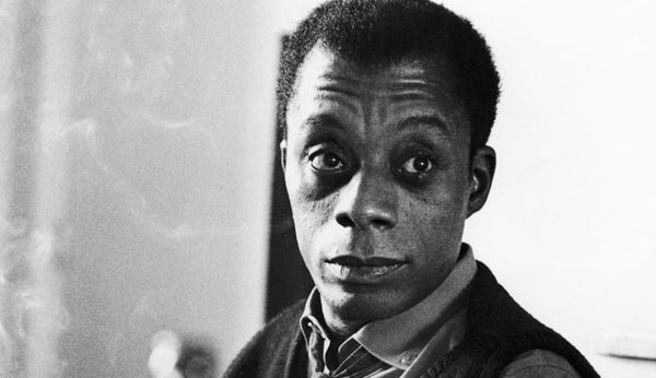 Photo, black and white, of James Baldwin looking past the camera, apparently wearing a collared shirt and sweater vest