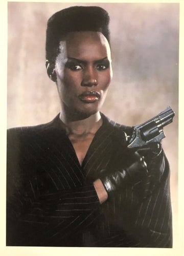 Grace is bosing in a black pinstripe suit while holding a gun. 