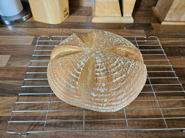 Round, golden loaf of white bread on a cooling rack. The loaf has circular white rings from the proving basket, and four slashes that are in the shape of a wonky cross.