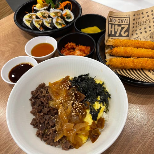 Stół z różnymi koreańskimi potrawami, w tym bibimbap, gimbap, dodatkami, smażoną krewetką zawiniętą w papier i sosami
