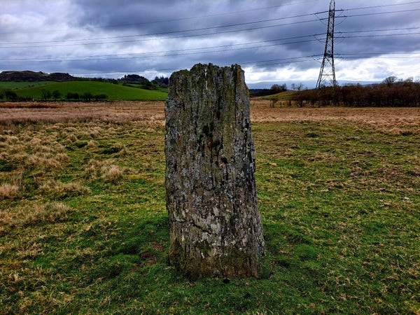 Coed-y-Bedo standing stone, a 6 foot slab of slate. Looking towards Y Berwyn mountains. A Pylon stands behind on the right. There is patchy cloud.