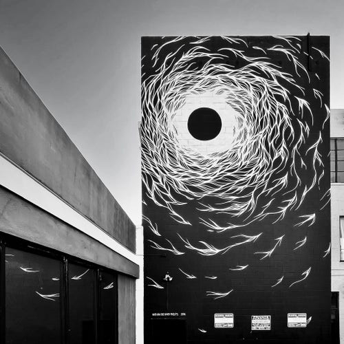 A striking black and white photograph of a beautiful and haunting 3-story mural on the side of a building. The mural centers around a large black dot that gives off the impression of being a celestial figure near the top, and all these angel or bird-like flourishes circling around floating upward and inward toward the dot. The background of the mural is deep black, but all the white swirling figures turn the background into a light color to that next to the dot the background it pure white. Big ‘eclipse’ vibes going on here. But I’ve also always thought of souls gently drifting to heaven as well. The image seems so peaceful and ethereal. The architecture surrounding the mural wall is very geometric and angular, and it helps draw the eye into the art. The glass windows on an adjacent building reflect the floating mural figures and when you look closely at the bottom of the mural you can see tiny No Parking signs to ground the image back to reality and give it a place in time. 