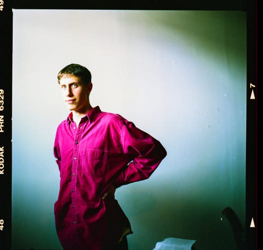 A tall, skinny young man with straight brown hair in a bowl cut stands in a shaft of light in front of a plain white wall. His hands rest on the small of his back, causing the oversized red button up shirt that he’s wearing to cinch at the waist. He looks piercingly and directly at the viewer.