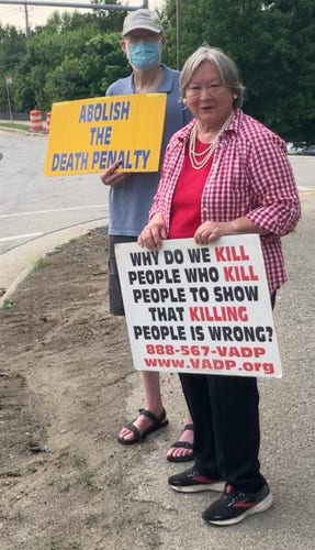 A man and a woman standing by a road holding signs and looking at the camera. The mans sign says "ABOLISH THE DEATH PENALTY" and the womans "WHY DO WE KILL PEOPLE WHO KILL PEOPLE TO SHOW THAT KILLING PEOPLE IS WRONG? 888-567-VADP WWW.VADP.org"