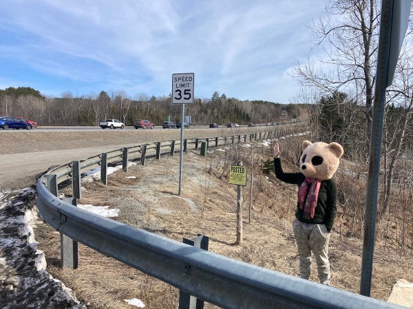 A woman wearing a sweater, camp pants and a pink scarf with a bear head on waves at busy traffic going by on a highway.