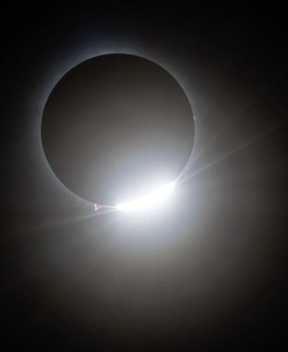 Photography. A color photo of a total solar eclipse of a total solar eclipse, seen in Vermont, USA on 08.04.2024. A black, round disk, behind which a thin, bright ring of rays appears. Below the moon, a white light hits the edge and illuminates the darkness with individual rays.