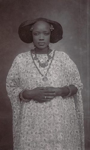 a black and white photo of a woman from her knees up, wearing a light dress with a print on it, her hair parted in a gorgeous shape, she wears two necklaces, has her hands folded over her abdomen, looking at the viewer, a grey toned background 