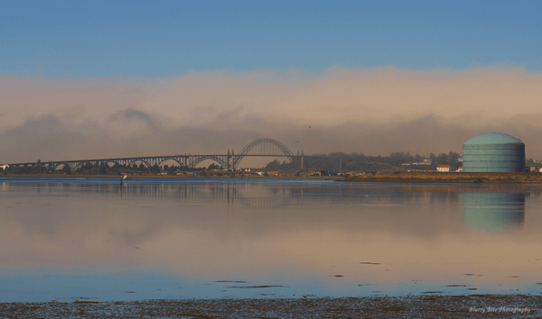 The Yaquina Bay Bridge appears in front of a receding fog bank, the image is mirrored by the calm waters on the bay in low tide