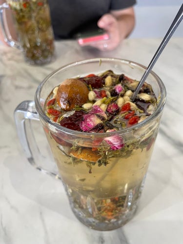 A glass mug filled with a tea that consists of various flower buds, tea leaves, and dried fruits.